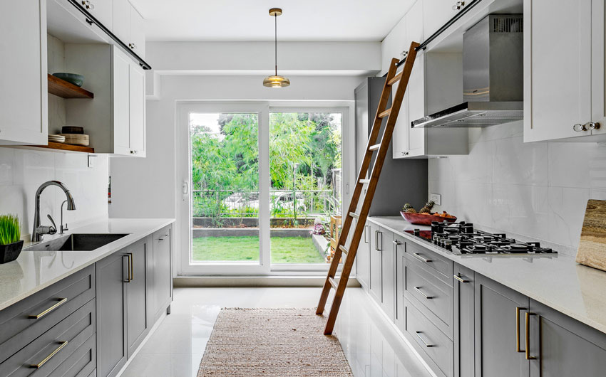 Galley kitchen with modern grey lower cabinets and clean white upper cabinets, high-end appliances, and a unique rolling ladder on a track to reach higher shelves. Large glass sliding doors on the far wall allowing lots of natural light.