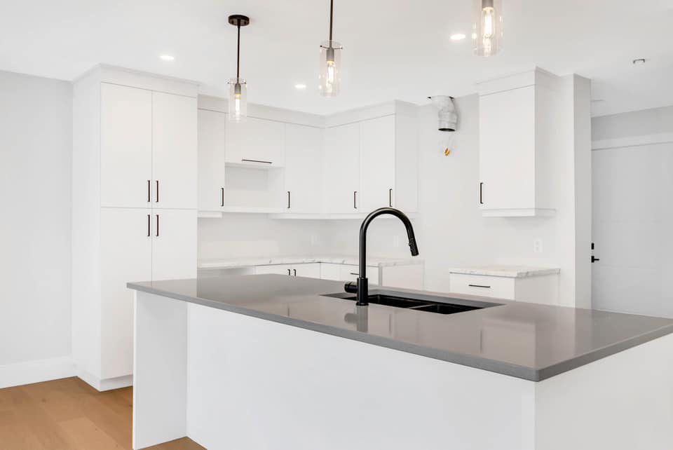 Directly looking at brand new modern all-white kitchen empty of appliances with a grey stone top island, with a black faucet, glass light fixtures above, and stainless steel hardware.